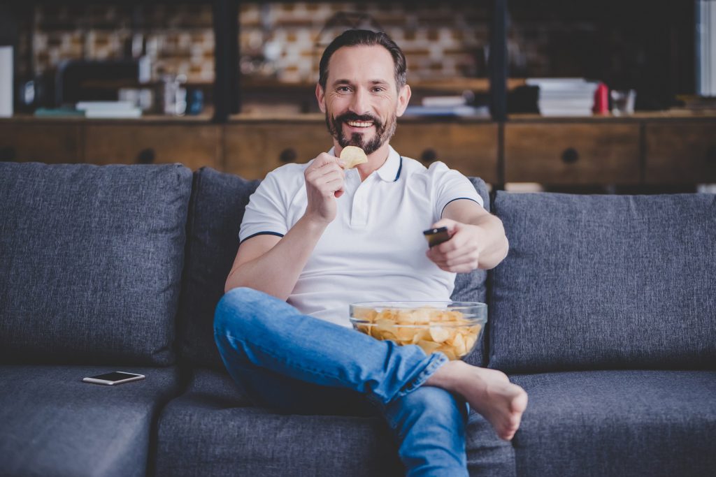 smiling man watching tv and eating chips at home