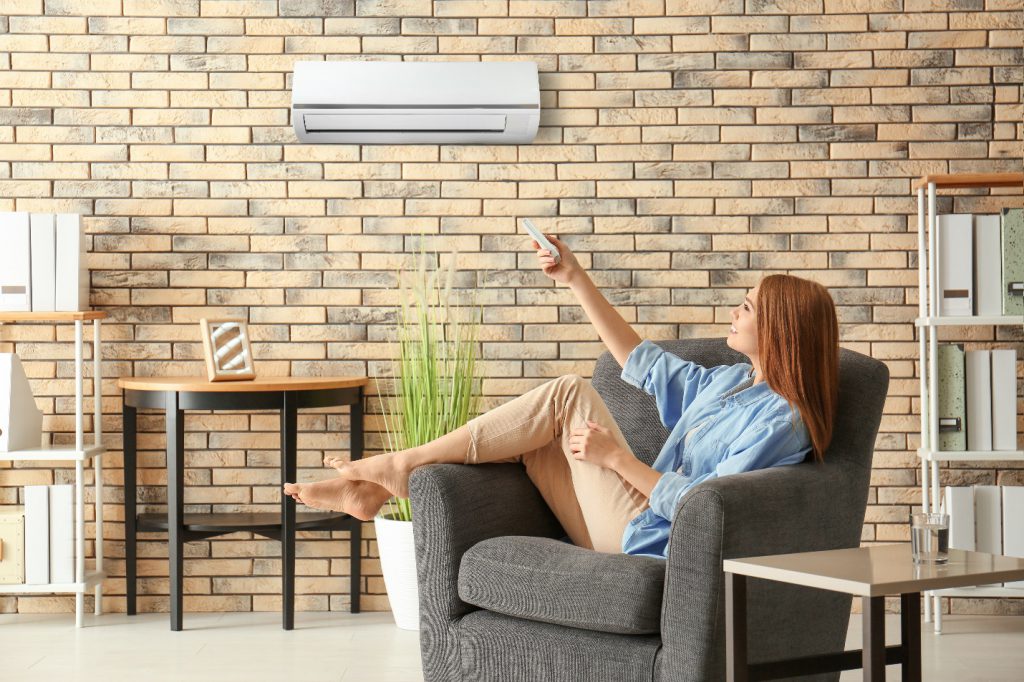 Young woman switching on air conditioner while sitting in armchair